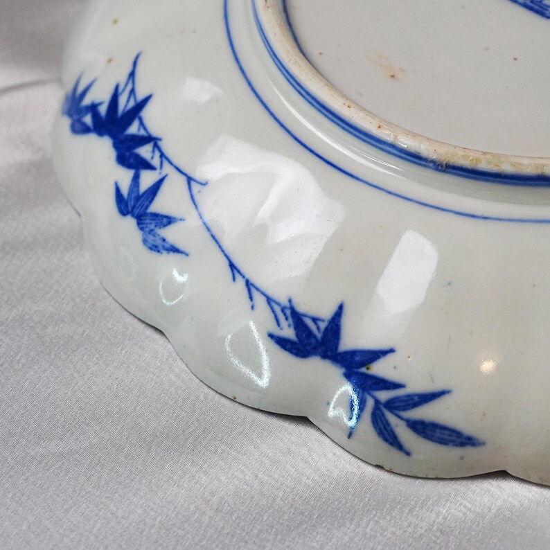 Puede incluir: Plato de cer&aacute;mica blanca con borde festoneado y dise&ntilde;o floral azul. El plato presenta un borde pintado de azul y un delicado patr&oacute;n de hojas y ramas azules. El plato es probablemente vintage o antiguo.