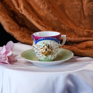 May include: A decorative teacup and saucer set. The cup features a white interior, a pink and blue rim, and gold floral accents. The saucer is light green. The set is placed on a white surface with a brown background.