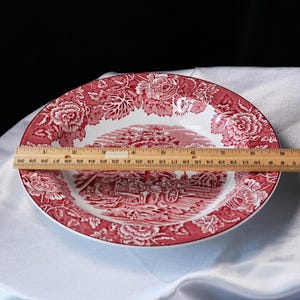 Op de afbeelding: Een ronde keramische bord met een rood-witte bloemenrand en een centraal landelijk tafereel met een paardenkoets. Een houten liniaal ligt over het midden en toont aan dat het bord een diameter heeft van ongeveer 23 cm.