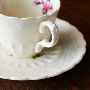 以下が含まれることがあります： 花柄の白いティーカップとソーサーのセット。ティーカップは曲線的なハンドルとスカラップの縁が付いています。ソーサーはマッチするスカラップの縁が付いています。このセットは磁器製で、繊細でヴィンテージな外観をしています。