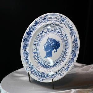 May include: White and blue commemorative plate featuring a profile of Queen Victoria. The plate has a blue floral border and text that reads "Jubilee Year" and "Compliments of Dei Gratia". The plate is on a metal stand.