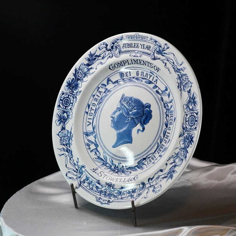 May include: White and blue commemorative plate featuring a profile of Queen Victoria. The plate has a blue floral border and text that reads "Jubilee Year" and "Compliments of Dei Gratia". The plate is on a metal stand.