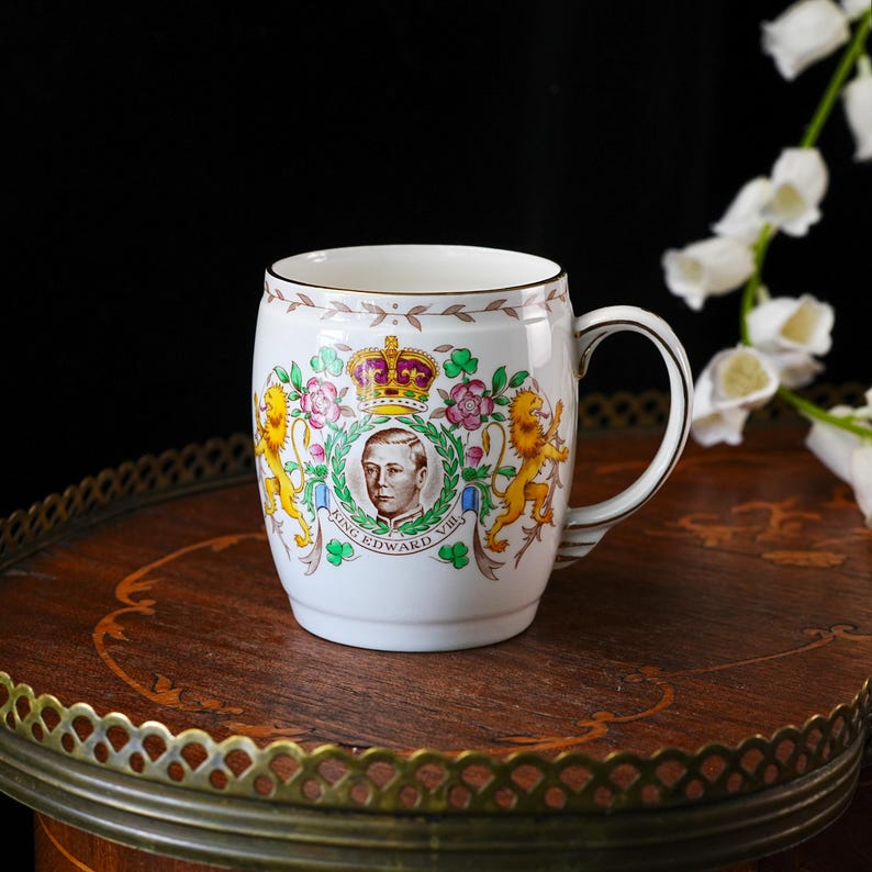 Może przedstawiać: Biały ceramiczny kubek z portretem kr&oacute;la Edwarda VIII umieszczonym wewnątrz zielonego wieńca laurowego. Wz&oacute;r zawiera kolorową koronę, dwa złote lwy, r&oacute;żowe r&oacute;że i zielone koniczyny. Poniżej znajduje się nadruk z tekstem KING EDWARD VIII.
