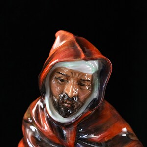 May include: A ceramic figure of a man in a red hooded robe. The figure has a beard and a thoughtful expression. The robe has dark red and black shading, and the hood has a white interior.