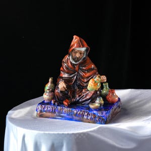 May include: A ceramic figurine of a robed figure with a hood, seated on a blue and brown base, surrounded by small, colourful bottles and vessels. The figurine has a detailed, hand-painted appearance, with a variety of colours.