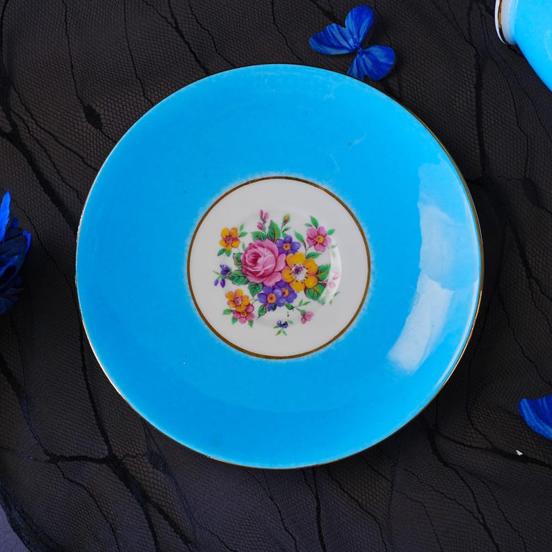 Op de afbeelding: Een ronde keramische schotel met een levendige turquoise blauwe rand en een wit cirkelvormig medaillon in het midden. Het centrum toont een gedetailleerd bloemstuk met roze, gele en paarse bloemen, afgewerkt met een dunne gouden rand.