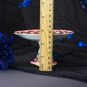 Puede incluir: Un plato de pedestal de porcelana rosa y blanca con patrones de filigrana dorada en el borde y la base. Una regla de madera est&aacute; colocada verticalmente contra el plato, mostrando una altura total de unos 8,3 cent&iacute;metros desde la base al borde.