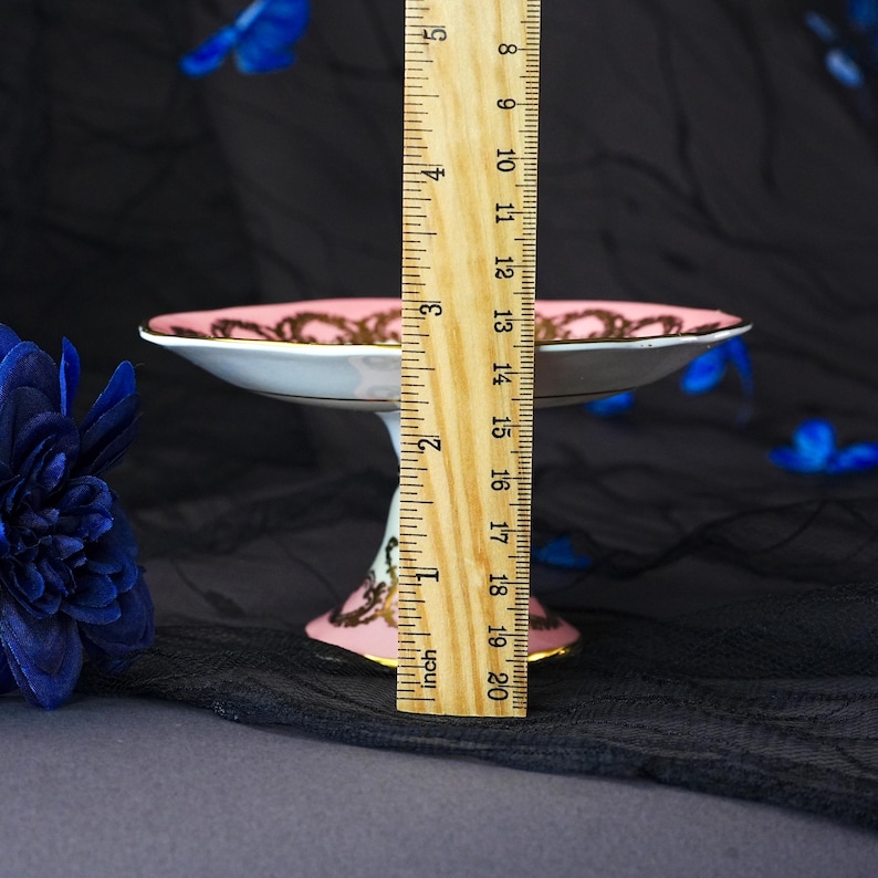 Puede incluir: Un plato de pedestal de porcelana rosa y blanca con patrones de filigrana dorada en el borde y la base. Una regla de madera est&aacute; colocada verticalmente contra el plato, mostrando una altura total de unos 8,3 cent&iacute;metros desde la base al borde.