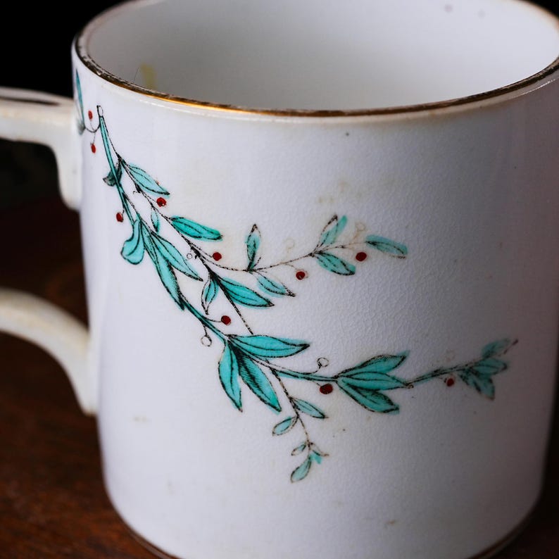May include: White ceramic mug with a gold rim and handle. The mug is decorated with a teal and red botanical design. The mug is a classic shape and appears to be vintage.