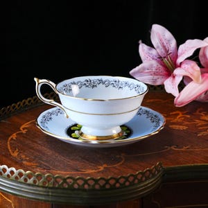 May include: A light blue porcelain teacup and saucer set featuring a delicate black floral pattern along the rims. The cup has a gold-trimmed handle and base, while the saucer displays a black circular accent with green leaf details.