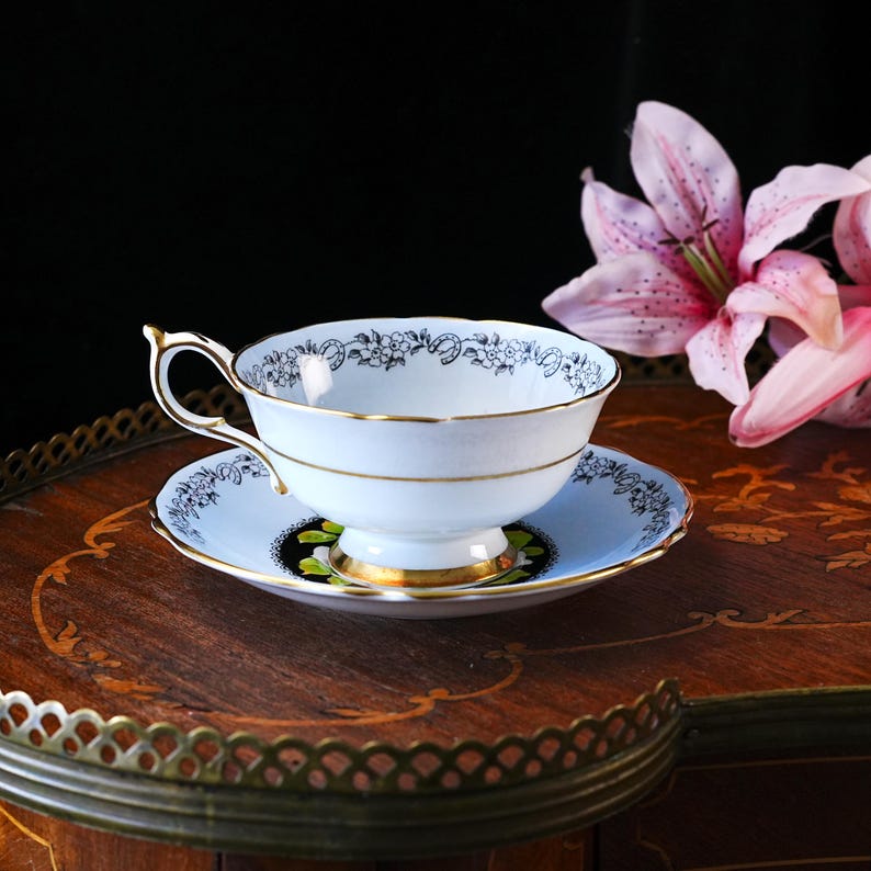 May include: A light blue porcelain teacup and saucer set featuring a delicate black floral pattern along the rims. The cup has a gold-trimmed handle and base, while the saucer displays a black circular accent with green leaf details.