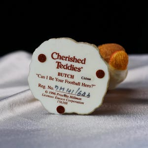 Op de afbeelding: Een "Cherished Teddies" beeldje met de tekst "Can I Be Your Football Hero?" en de naam "BUTCH". De basis is gebroken wit met bruine accenten en tekst. Het beeldje is gemaakt in China.