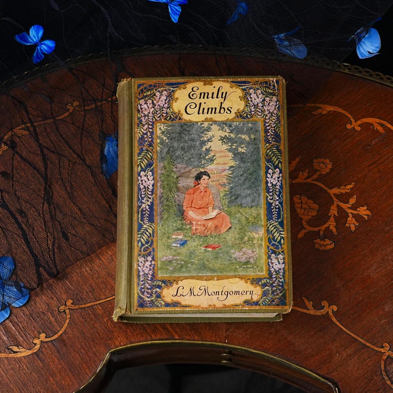 Puede incluir: Un libro de tapa dura vintage titulado Emily Climbs de L.M. Montgomery. La portada presenta una ilustraci&oacute;n central de una persona con un vestido rojo sentada en una zona de hierba rodeada de &aacute;rboles, enmarcada por un borde decorativo de flores moradas y oro.