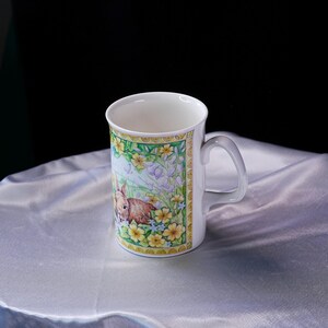 Puede incluir: Una taza de cer&aacute;mica blanca con una ilustraci&oacute;n colorida de un conejo marr&oacute;n rodeado de flores amarillas y campanillas azules. El dise&ntilde;o est&aacute; enmarcado por un borde decorativo y la taza incluye un asa curva en el lado derecho.