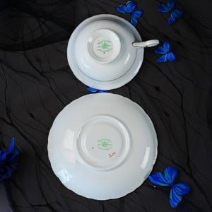 K&ouml;nnte beinhalten: Ein wei&szlig;es Porzellan-Tassenset mit Untertasse von Royal Grafton, von der Unterseite gezeigt. Die Teile zeigen ein gr&uuml;nes Kronenlogo und den Text Royal Grafton Made in England. Die Tasse hat einen goldverzierten Henkel und einen gewellten Rand.