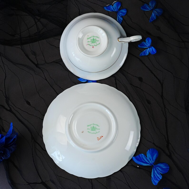 K&ouml;nnte beinhalten: Ein wei&szlig;es Porzellan-Tassenset mit Untertasse von Royal Grafton, von der Unterseite gezeigt. Die Teile zeigen ein gr&uuml;nes Kronenlogo und den Text Royal Grafton Made in England. Die Tasse hat einen goldverzierten Henkel und einen gewellten Rand.