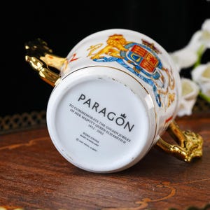 Puede incluir: Una taza de porcelana blanca con asas doradas, con un escudo real y el texto en la base: PARAGON, TO COMMEMORATE THE GOLDEN JUBILEE OF HER MAJESTY QUEEN ELIZABETH II 1952-2002, BONE CHINA, MADE IN ENGLAND, 2001 ROYAL ALBERT.