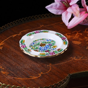 Puede incluir: Un peque&ntilde;o plato de cer&aacute;mica blanca con borde dorado festoneado, que presenta un borde floral colorido y un emblema central con una corona y el texto ER Coronation 2nd June 1953. El dise&ntilde;o incluye hojas verdes y flores de colores.