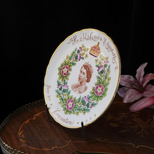 Op de afbeelding: Een wit herdenkingsbord van porselein met een gouden rand, voorzien van een portret van koningin Elizabeth II. Het ontwerp bevat een kroon, een bloemenkrans met rozen en distels, en de tekst H.M. Queen Elizabeth II, June 2nd, Coronation 1953.