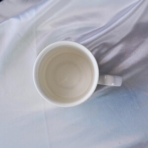 Puede incluir: Una taza de cer&aacute;mica blanca lisa vista directamente desde arriba, mostrando la abertura circular y la base interior. Un asa &uacute;nica se extiende desde el lado derecho de la taza, la cual est&aacute; colocada sobre una superficie de tela lisa y clara.
