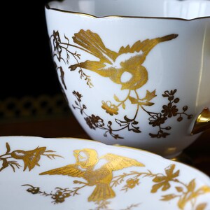Op de afbeelding: Witte theekop en schotel set met gouden vogel- en bloemmotieven. De theekop heeft een geschulpte rand en een gouden oor. De schotel heeft ook een gouden vogel- en bloemmotief. De set is waarschijnlijk van porselein of keramiek.