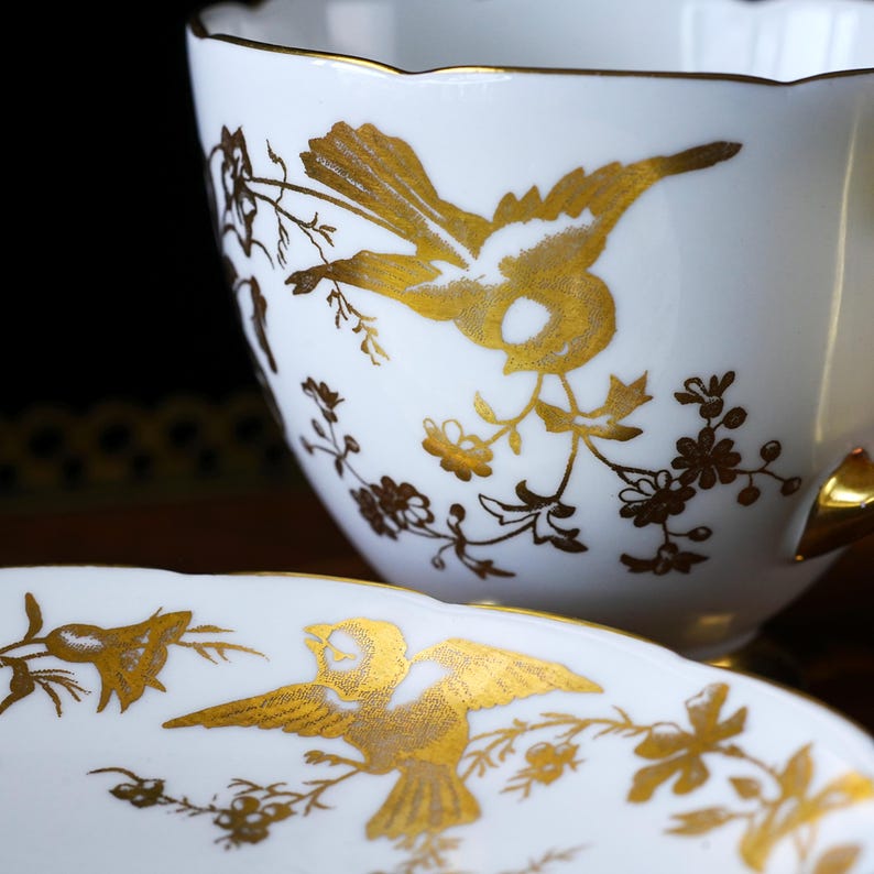 Op de afbeelding: Witte theekop en schotel set met gouden vogel- en bloemmotieven. De theekop heeft een geschulpte rand en een gouden oor. De schotel heeft ook een gouden vogel- en bloemmotief. De set is waarschijnlijk van porselein of keramiek.