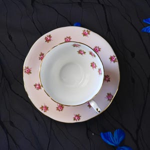 Może przedstawiać: Zestaw filiżanki do herbaty ze spodkiem z jasnor&oacute;żowej porcelany, ozdobiony powtarzającym się wzorem małych r&oacute;żowych r&oacute;ż z zielonymi liśćmi. Filiżanka i spodek wykończone są cienką złotą obw&oacute;dką na krawędziach. Wnętrze filiżanki jest białe.