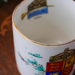May include: A white ceramic mug with a gold rim and a detailed crest design. The crest features a crown, a banner, and a shield with red, blue, and yellow elements. The mug has a cylindrical shape and appears to be vintage.