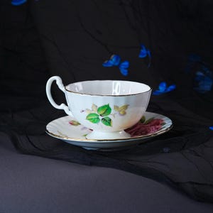 Peut inclure: Un ensemble tasse et soucoupe en porcelaine blanche orn&eacute; d'un motif floral d&eacute;licat avec des feuilles vertes et une rose rose. La tasse pr&eacute;sente un bord festonn&eacute; et une bordure dor&eacute;e, accompagn&eacute;e d'une soucoupe assortie avec une rose rose.