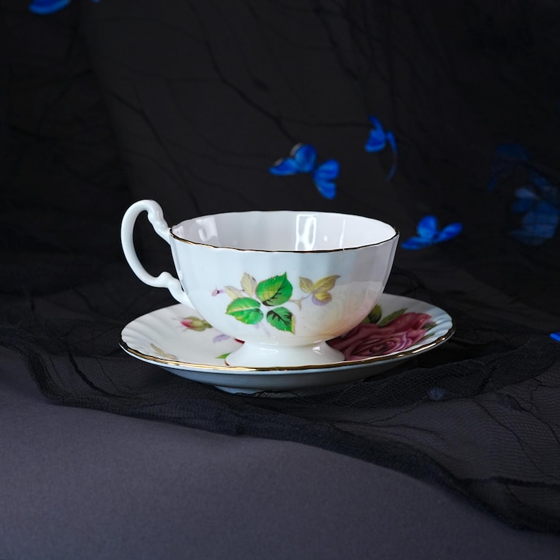 Peut inclure: Un ensemble tasse et soucoupe en porcelaine blanche orn&eacute; d'un motif floral d&eacute;licat avec des feuilles vertes et une rose rose. La tasse pr&eacute;sente un bord festonn&eacute; et une bordure dor&eacute;e, accompagn&eacute;e d'une soucoupe assortie avec une rose rose.