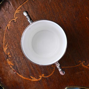 May include: A white teacup with silver trim and handles, viewed from above. The cup rests on a dark wooden surface with inlaid decoration. The cup's interior is a bright white, contrasting with the silver and dark wood.