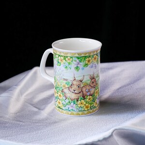 Puede incluir: Una taza de cer&aacute;mica blanca con una colorida ilustraci&oacute;n de dos conejos marrones rodeados de flores amarillas y azul claro. El dise&ntilde;o incluye un borde decorativo en la parte superior e inferior, con un asa lateral y un acabado liso y brillante.