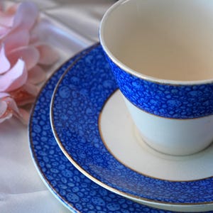 May include: A white teacup and saucer set with a blue bubble pattern and gold trim. The set includes a small plate, a saucer, and a teacup. The teacup is white with a blue band around the top.
