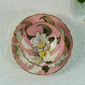 May include: A pink teacup and saucer set with a floral design. The teacup and saucer are decorated with a large white and yellow flower with green leaves. The rims are trimmed with gold.