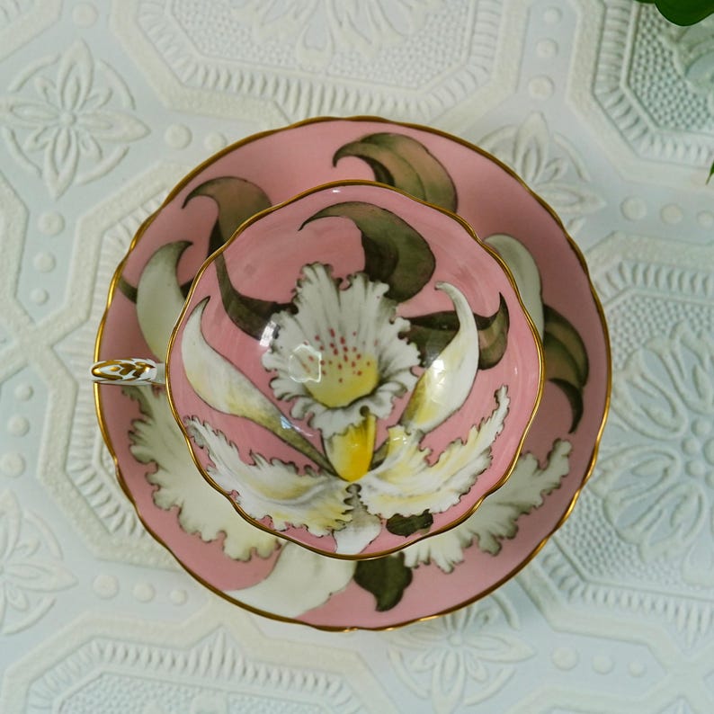 May include: A pink teacup and saucer set with a floral design. The teacup and saucer are decorated with a large white and yellow flower with green leaves. The rims are trimmed with gold.