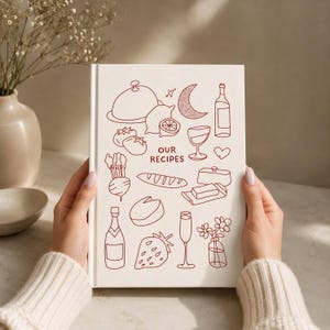 May include: A white hardcover recipe book featuring dark red line art illustrations of food and drink items, including a cloche, lemon, wine bottle, glass, heart, bread, butter, cheese, strawberry, and flowers, with the text OUR RECIPES centered.