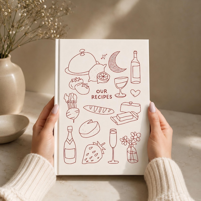 May include: A white hardcover recipe book featuring dark red line art illustrations of food and drink items, including a cloche, lemon, wine bottle, glass, heart, bread, butter, cheese, strawberry, and flowers, with the text OUR RECIPES centered.