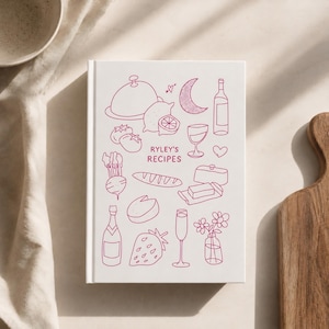 May include: A white hardcover recipe book titled Ryley's Recipes in pink ink. The cover features line art illustrations of food and drink items including a cloche, lemon, crescent moon, wine bottle, glass, heart, radish, bread, butter, cheese, strawberry, and flowers.