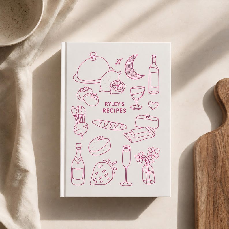 May include: A white hardcover recipe book titled Ryley's Recipes in pink ink. The cover features line art illustrations of food and drink items including a cloche, lemon, crescent moon, wine bottle, glass, heart, radish, bread, butter, cheese, strawberry, and flowers.
