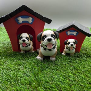 Op de afbeelding: Drie bruine en witte bulldog-figuurtjes, elk staand voor een rood en zwart hondenhok met een wit bot op het dak. De hondenhokken zijn van verschillende grootte, en de figuurtjes kijken allemaal naar voren met hun tongen uitgestoken.