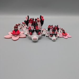 May include: Three black and white 3D printed figurines with pink and red flower accents. The figurines are in the shape of a stylized animal with large ears and a long tail.
