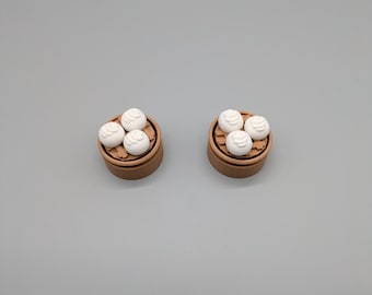 May include: Two miniature dim sum food charms. Each charm features a brown bamboo steamer basket with three white dumplings. The dumplings have a textured surface. The charms are set against a gray background.