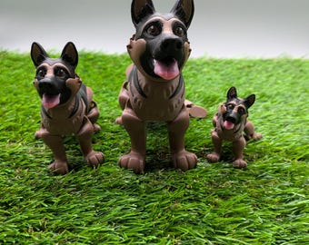 May include: Three German Shepherd dog figurines in different sizes, standing on a green artificial grass surface. The largest figurine is brown and black with a pink tongue sticking out. The medium-sized figurine is brown and black with a pink tongue sticking out. The smallest figurine is brown and black with a pink tongue sticking out.