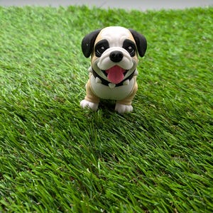 Op de afbeelding: Een klein, beige en wit bulldog-beeldje met zwarte oren en een zwarte neus. Het beeldje glimlacht met zijn tong naar buiten en heeft een zwarte halsband. Het beeldje zit op een groene kunstgras ondergrond.