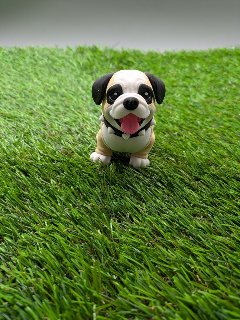 Op de afbeelding: Een klein, beige en wit bulldog-beeldje met zwarte oren en een zwarte neus. Het beeldje glimlacht met zijn tong naar buiten en heeft een zwarte halsband. Het beeldje zit op een groene kunstgras ondergrond.