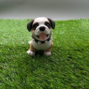 Op de afbeelding: Een klein, bruin en wit bulldog-beeldje met zwarte oren en een zwarte halsband met zilveren studs. Het beeldje staat op een groene kunstgras ondergrond.