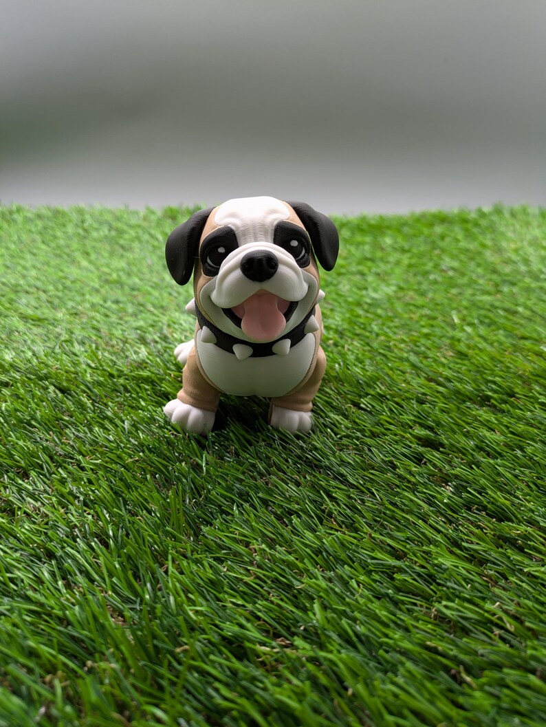 Op de afbeelding: Een klein, bruin en wit bulldog-beeldje met zwarte oren en een zwarte halsband met zilveren studs. Het beeldje staat op een groene kunstgras ondergrond.
