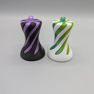 May include: Two decorative spinning tops with a twisted, hourglass-like silhouette. One top features a matte black base with metallic purple spiral accents. The second top has a matte white base with metallic green spiral accents on the surface.