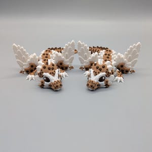 May include: Two whimsical, spotted dragon figurines with white wings and a brown and white color scheme. The dragons have detailed features, including small claws and expressive eyes. The figurines are displayed on a plain gray background.