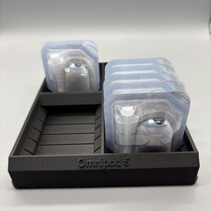 May include: A black rectangular tray with the text "Omnipod 5" holds several sealed, clear plastic packages. Each package contains a medical device. The tray has multiple compartments.